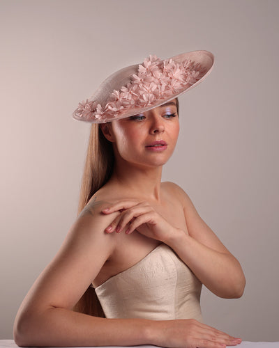 Model is showcasing the front right side of the medium size pink saucer hat, curved towards the left side and tilted to the right side, is decorated with multi petal pink silk sateen flowers, from top left side to right side. Centre of the flowers are embellished with pink pearls.