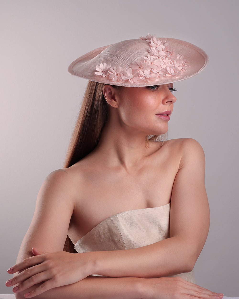 Model is showcasing the front from the right angle of the medium size pink saucer hat, curved towards the left side and tilted to the right side, is decorated with multi petal pink silk sateen flowers, from top left side to right side. Centre of the flowers are embellished with pink pearls.