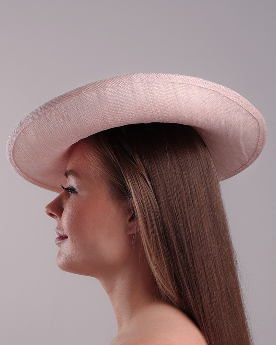 Model is showcasing the left side of the medium size pink saucer hat, curved towards the left side and tilted to the right side, is decorated with multi petal pink silk sateen flowers, from top left side to right side. Centre of the flowers are embellished with pink pearls.