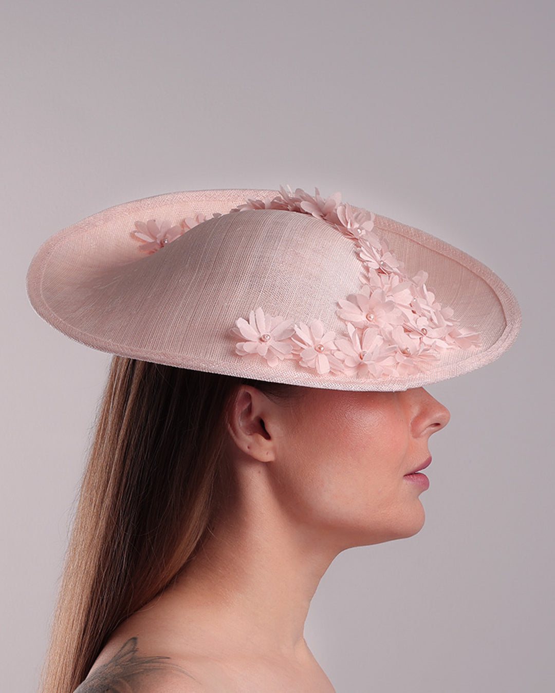 Model is showcasing the right side of the medium size pink saucer hat, curved towards the left side and tilted to the right side, is decorated with multi petal pink silk sateen flowers, from top left side to right side. Centre of the flowers are embellished with pink pearls.