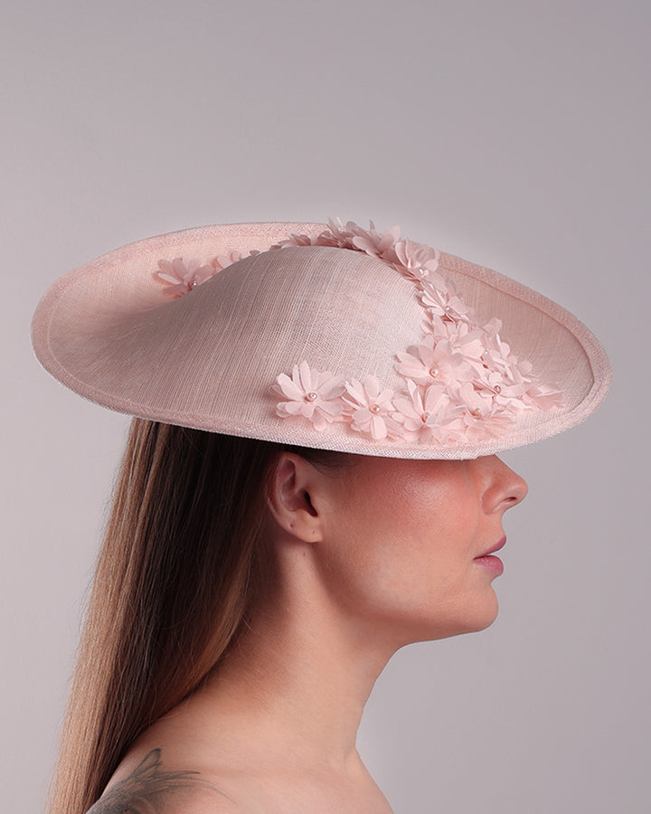 Model is showcasing the right side of the medium size pink saucer hat, curved towards the left side and tilted to the right side, is decorated with multi petal pink silk sateen flowers, from top left side to right side. Centre of the flowers are embellished with pink pearls.