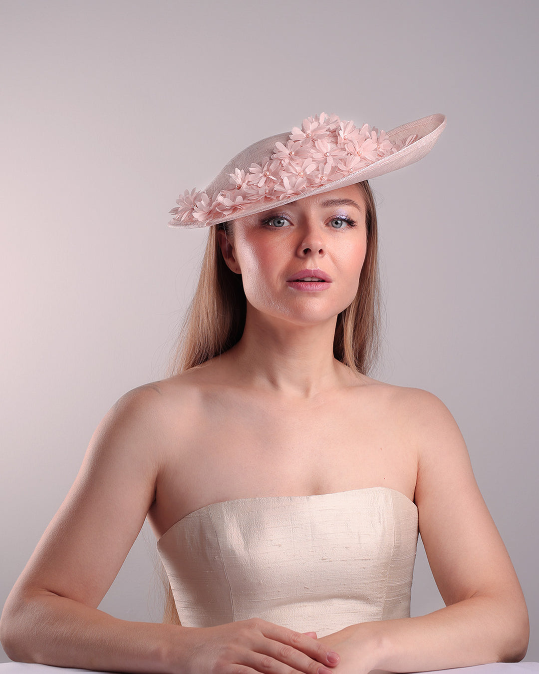 Model is showcasing the front of the medium size pink saucer hat, curved towards the left side and tilted to the right side, is decorated with multi petal pink silk sateen flowers, from top left side to right side. Centre of the flowers are embellished with pink pearls.