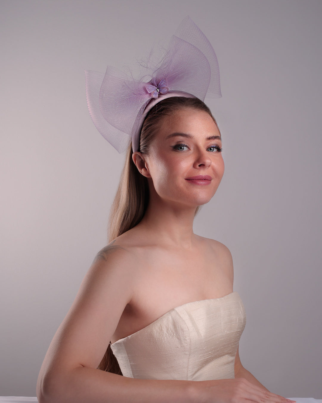 The model is showcasing the front right side of a semi high dark lilac silk headband decorated with double layerd large crinoline bow embellished with a silk organza butterfly.