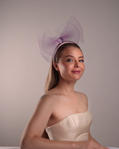 The model is showcasing the front right side of a semi high dark lilac silk headband decorated with double layerd large crinoline bow embellished with a silk organza butterfly.