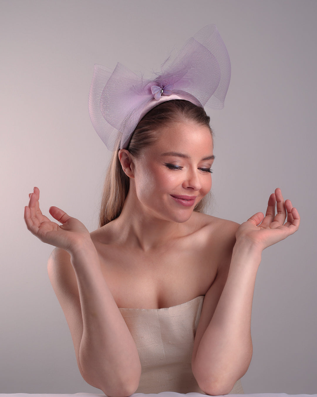 The model is showcasing the front top side of a semi high dark lilac silk headband decorated with double layerd large crinoline bow embellished with a silk organza butterfly.