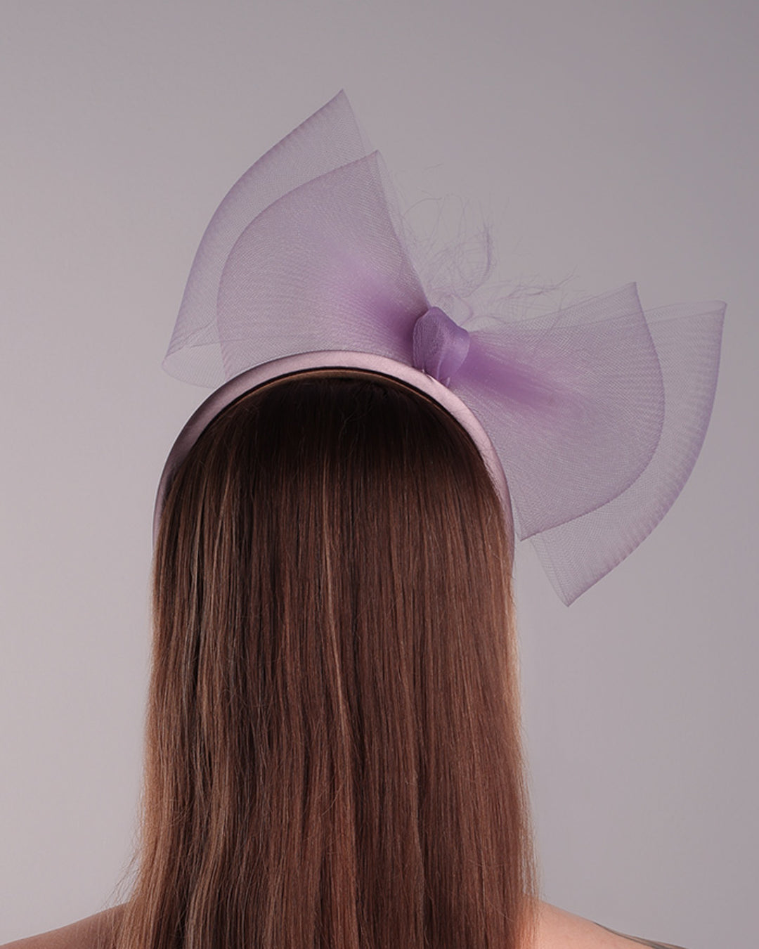 The model is showcasing the back side of a semi high dark lilac silk headband decorated with double layerd large crinoline bow embellished with a silk organza butter