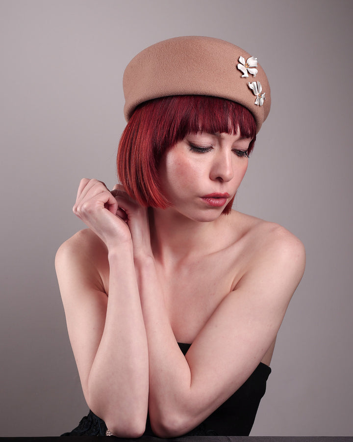 The female model is wearing and showcasing the left angle front side of a camel-coloured felt beret with matte white-coloured metal flowers. The beret has a layer on the right side which the wearer also tilts towards.. Designed by Merve Bayindir.