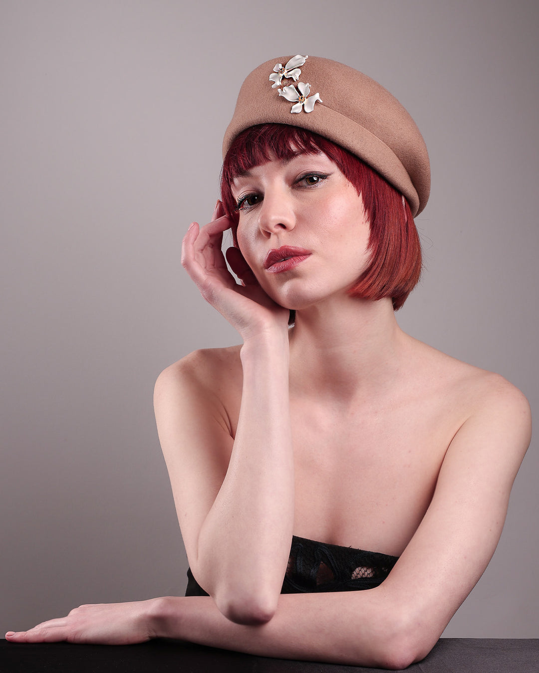The female model is wearing and showcasing the right angle front side of a camel-coloured felt beret with matte white-coloured metal flowers. The beret has a layer on the right side which the wearer also tilts towards.. Designed by Merve Bayindir.