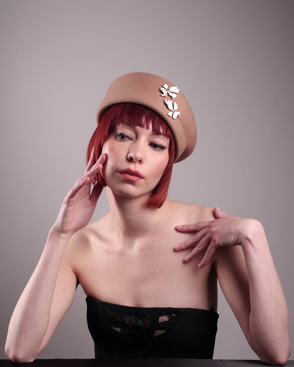 The female model is wearing and showcasing the front side of a camel-coloured felt beret with matte white-coloured metal flowers. The beret has a layer on the right side which the wearer also tilts towards.. Designed by Merve Bayindir.