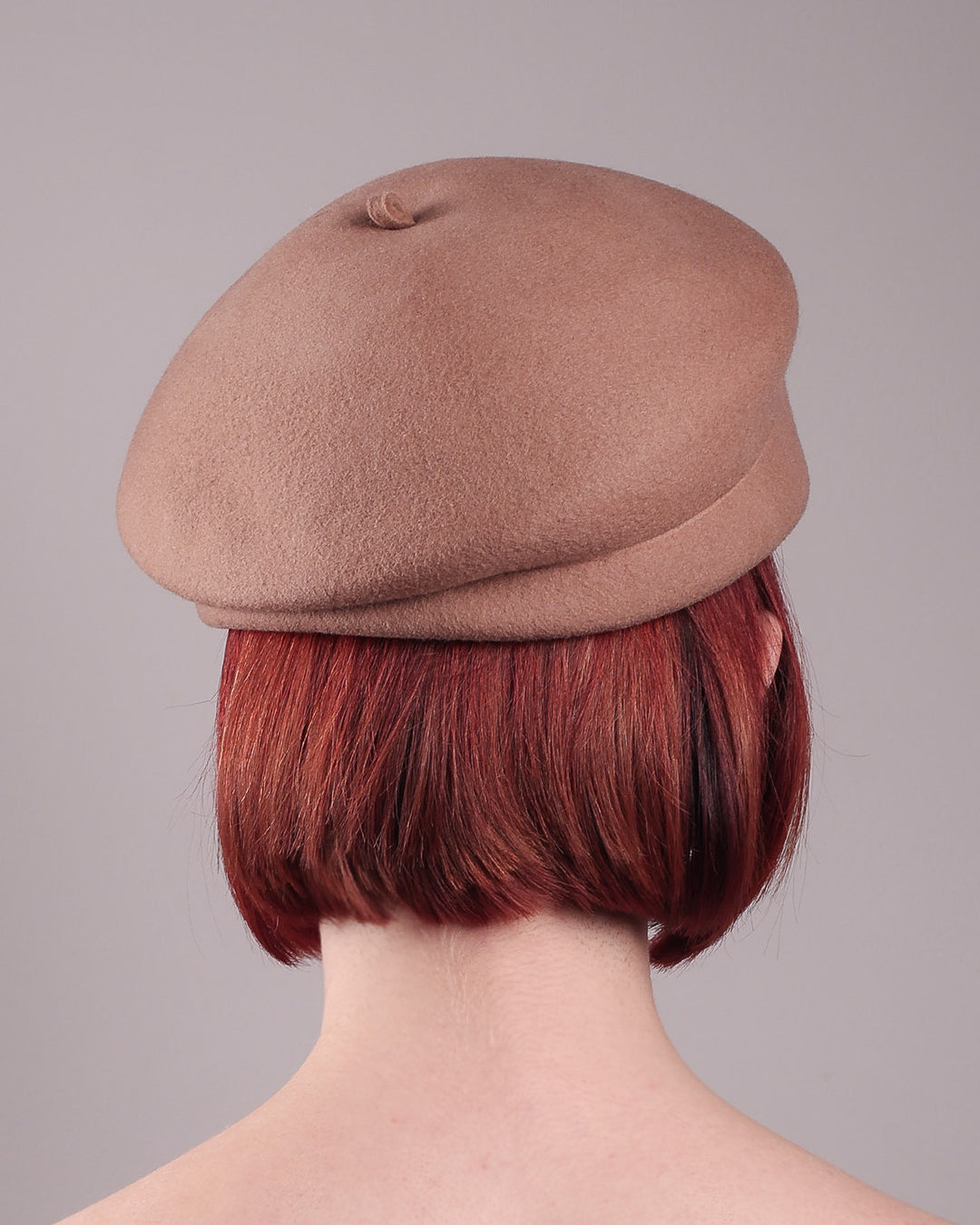 The female model is wearing and showcasing theback of a camel-coloured felt beret with matte white-coloured metal flowers. The beret has a layer on the right side which the wearer also tilts towards.. Designed by Merve Bayindir.