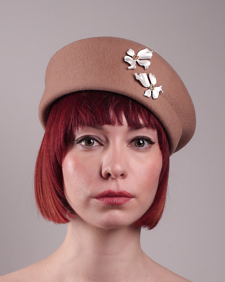 The female model is wearing and showcasing the front close up of a camel-coloured felt beret with matte white-coloured metal flowers. The beret has a layer on the right side which the wearer also tilts towards.. Designed by Merve Bayindir.