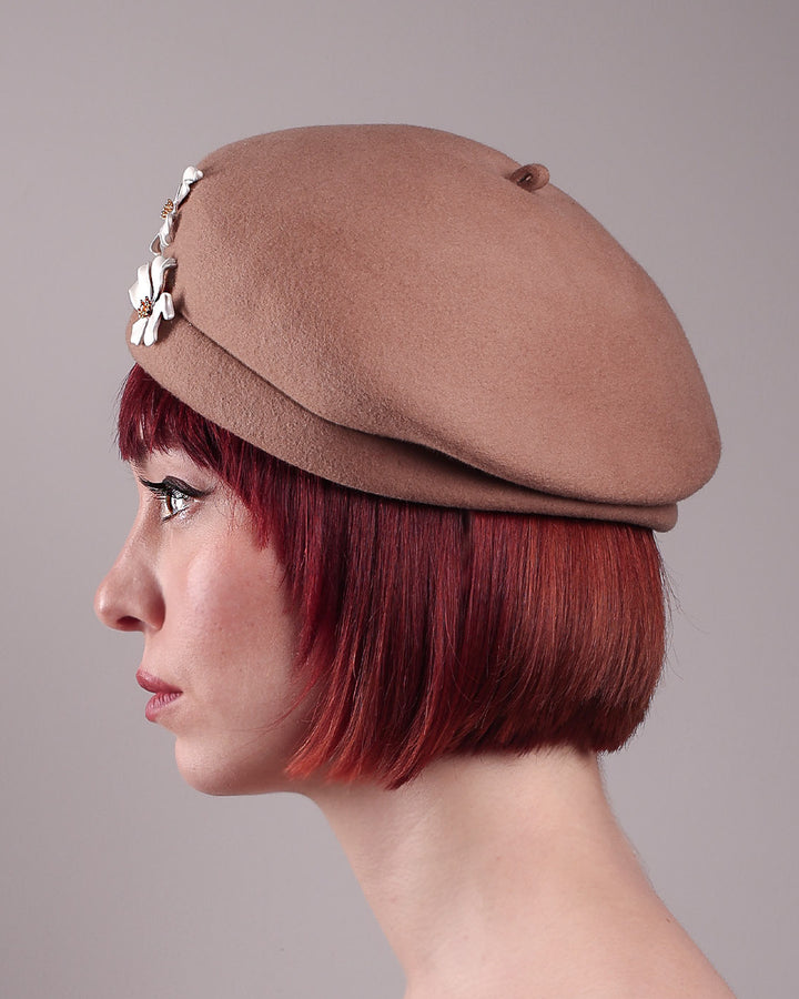 The female model is wearing and showcasing the left side close up of a camel-coloured felt beret with matte white-coloured metal flowers. The beret has a layer on the right side which the wearer also tilts towards.. Designed by Merve Bayindir.