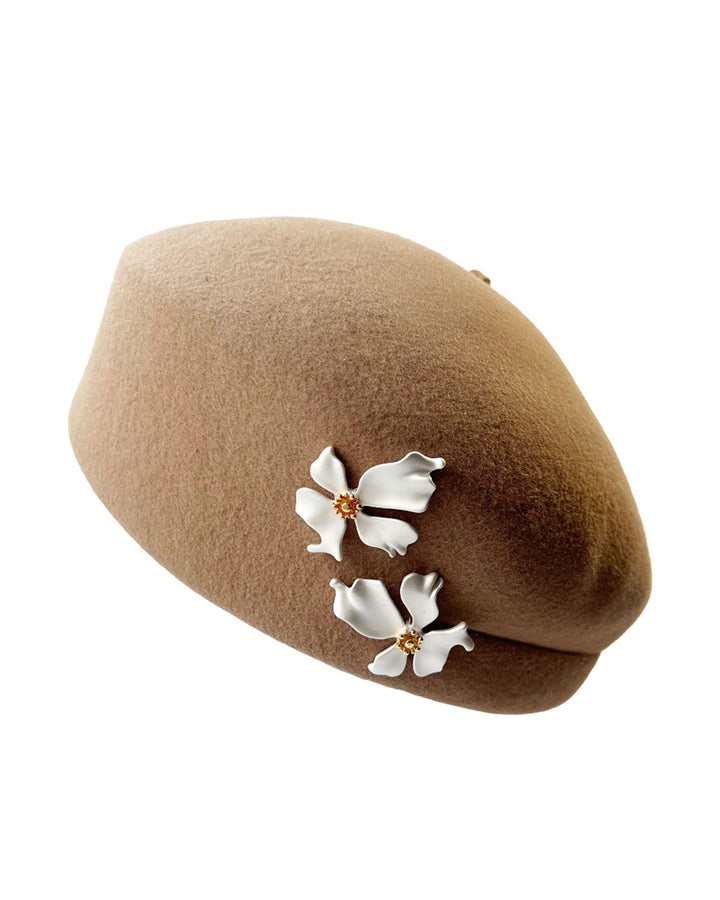 The still life picture of camel-coloured felt beret with matte white-coloured metal flowers. The beret has a layer on the right side which the wearer also tilts towards. Designed by Merve Bayindir.
