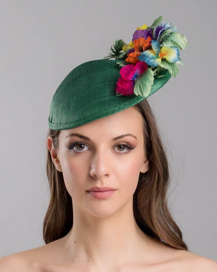 Model is showcasing the front side close up of small, asymmetric emerald green saucer hat decorated with multi coloured violets on the left. Created and designed by Merve bayindir millinery.