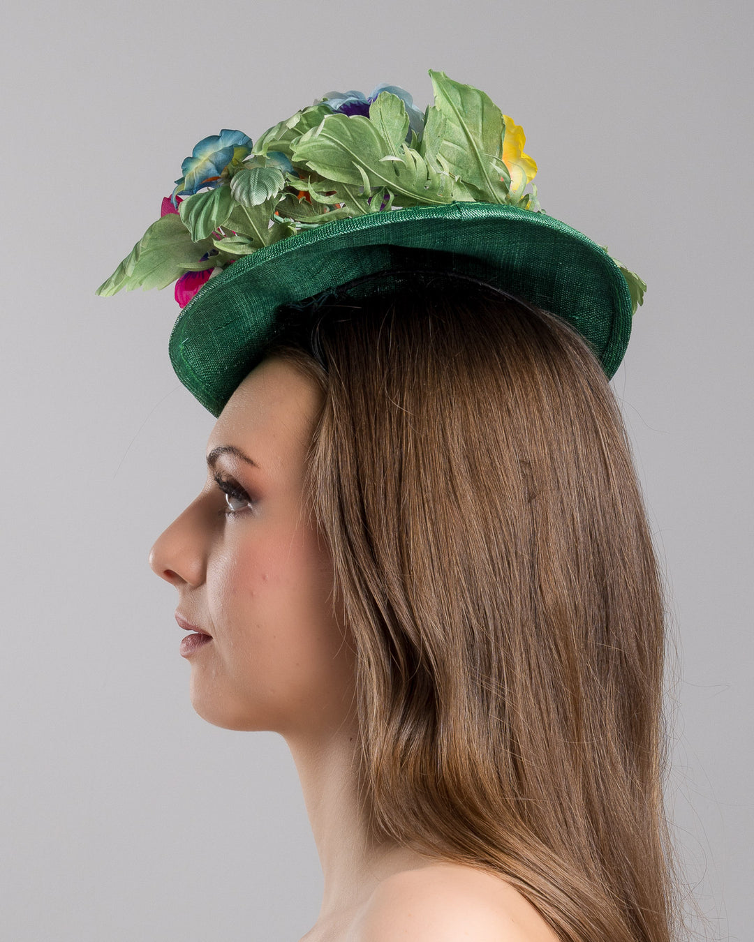 Model is showcasing the left side close up of small, asymmetric emerald green saucer hat decorated with multi coloured violets on the left. Created and designed by Merve bayindir millinery.