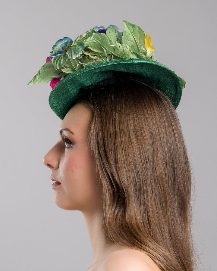 Model is showcasing the left side close up of small, asymmetric emerald green saucer hat decorated with multi coloured violets on the left. Created and designed by Merve bayindir millinery.