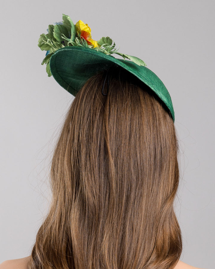 Model is showcasing the back side close up of small, asymmetric emerald green saucer hat decorated with multi coloured violets on the left. Created and designed by Merve bayindir millinery.