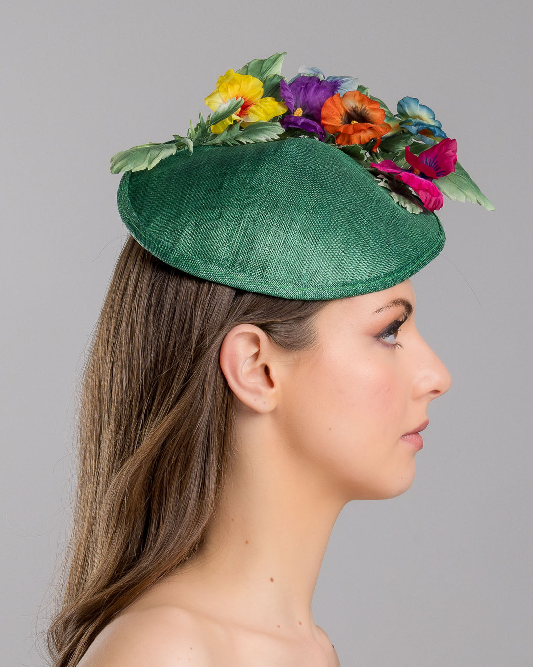 Model is showcasing the right side close up of small, asymmetric emerald green saucer hat decorated with multi coloured violets on the left. Created and designed by Merve bayindir millinery.