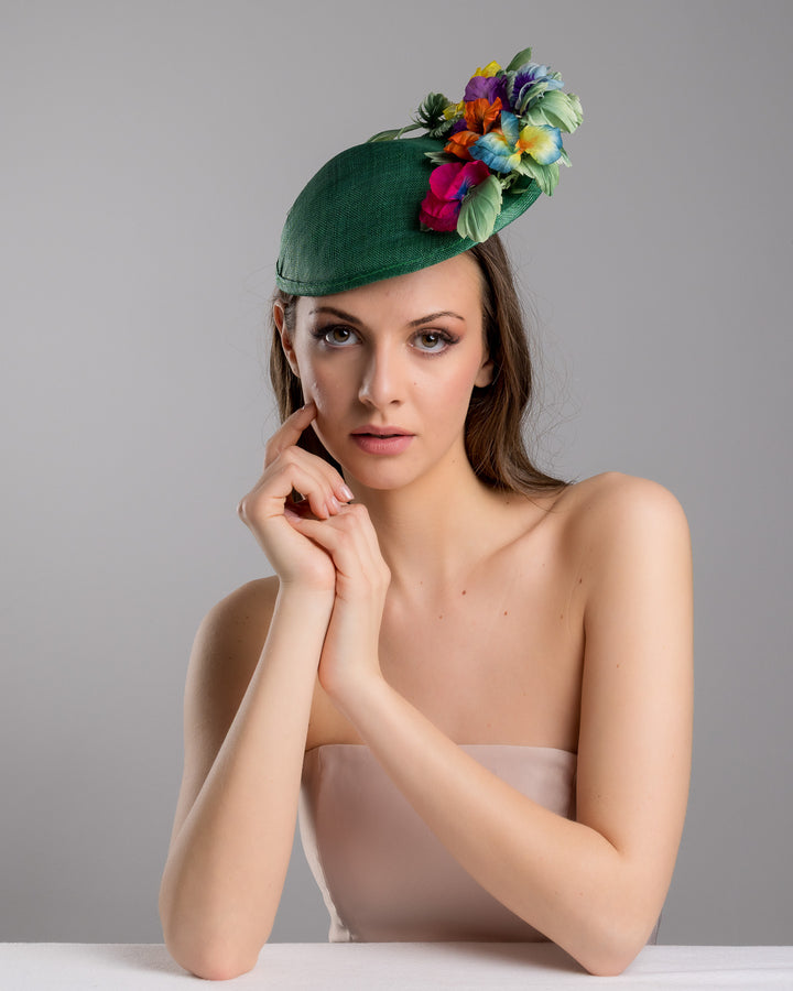 Female model is posing wearing the Peyton a small, asymmetric emerald green saucer hat decorated with multi coloured violets on the left. Created and designed by Merve bayindir millinery.