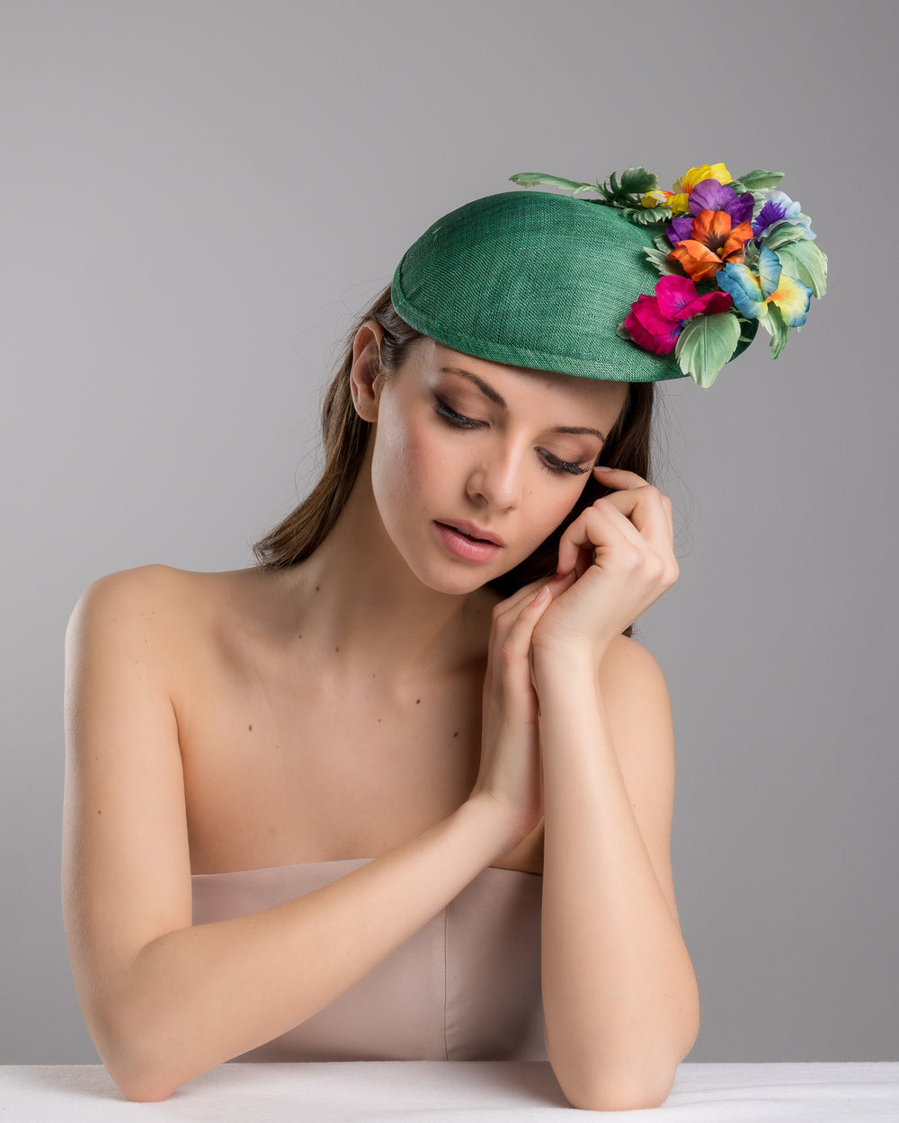 Female model is posing wearing the Peyton a small, asymmetric emerald green saucer hat decorated with multi coloured violets on the left. Created and designed by Merve bayindir millinery.