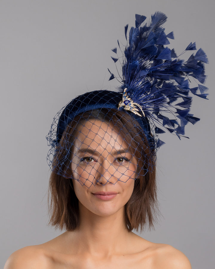 Model is showcasing the front side close up of navy coloured high velvet headband, covering half of the face with a stiffened navy veil elevated with a gold coloured metal bird with a large navy rhinestone and navy feathers behind the word placed high and like a fan. Made by Merve Bayindir Millinery.
