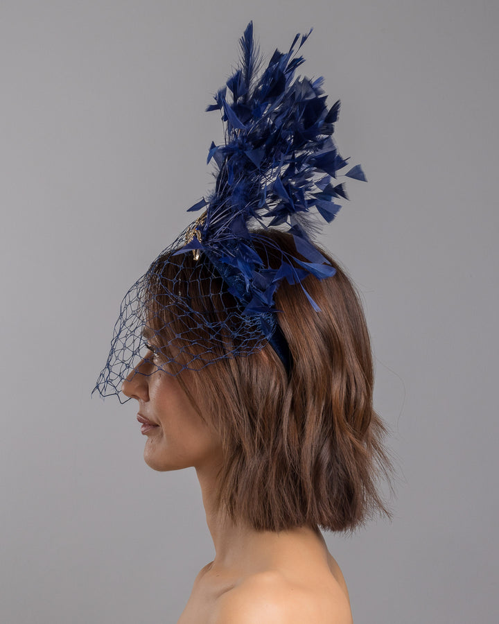 Model is showcasing the left side close up of navy coloured high velvet headband, covering half of the face with a stiffened navy veil elevated with a gold coloured metal bird with a large navy rhinestone and navy feathers behind the word placed high and like a fan. Made by Merve Bayindir Millinery.