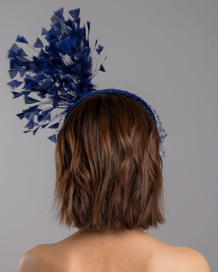 Model is showcasing the back side close up of navy coloured high velvet headband, covering half of the face with a stiffened navy veil elevated with a gold coloured metal bird with a large navy rhinestone and navy feathers behind the word placed high and like a fan. Made by Merve Bayindir Millinery.