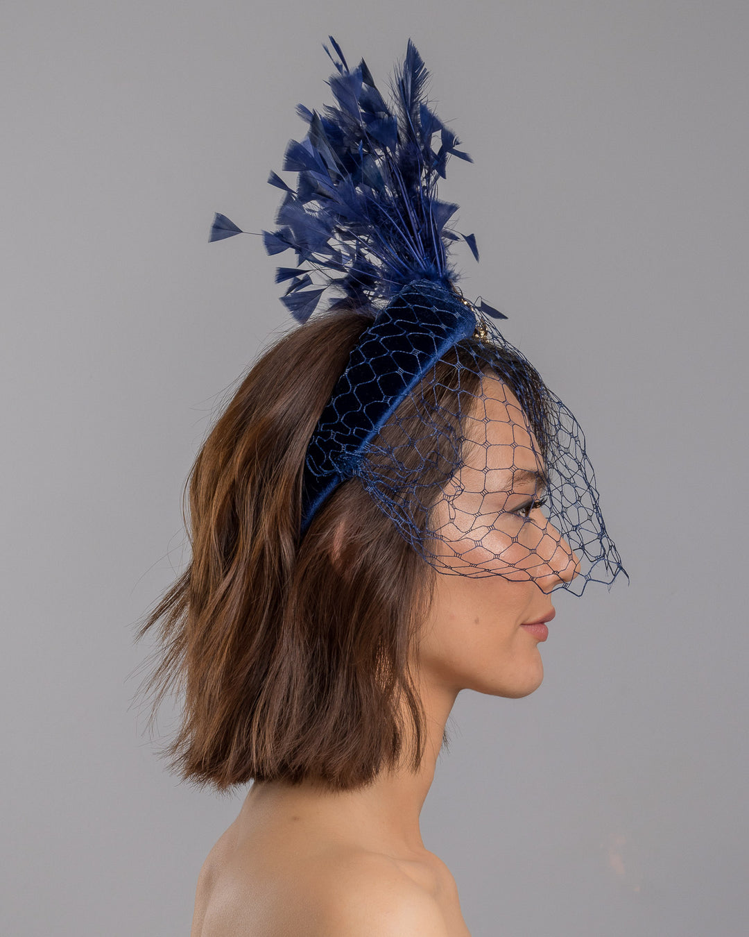 Model is showcasing the right side close up of navy coloured high velvet headband, covering half of the face with a stiffened navy veil elevated with a gold coloured metal bird with a large navy rhinestone and navy feathers behind the word placed high and like a fan. Made by Merve Bayindir Millinery.