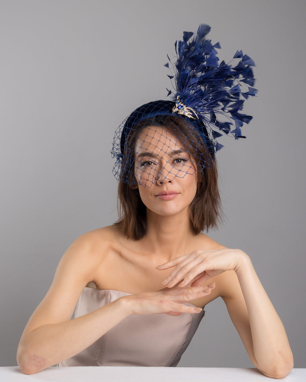 Female model is posing wearing Phoenix the navy coloured high velvet headband, covering half of the face with a stiffened navy veil elevated with a gold coloured metal bird with a large navy rhinestone and navy feathers behind the word placed high and like a fan. Made by Merve Bayindir Millinery.