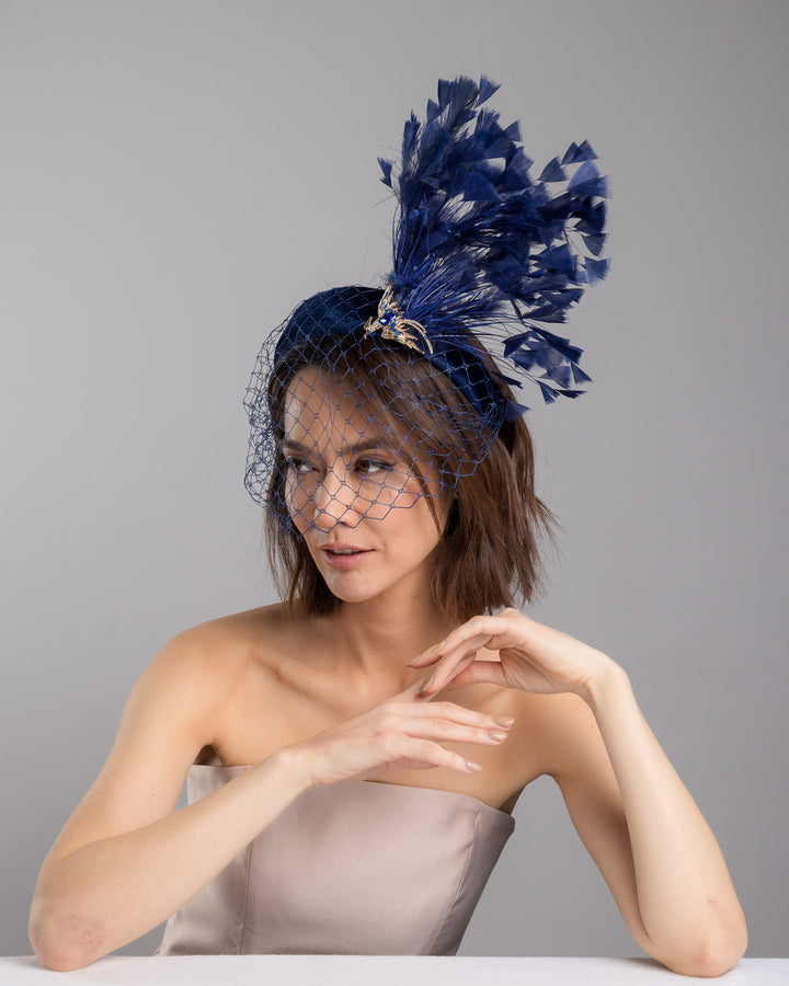 Female model is posing wearing Phoenix the navy coloured high velvet headband, covering half of the face with a stiffened navy veil elevated with a gold coloured metal bird with a large navy rhinestone and navy feathers behind the word placed high and like a fan. Made by Merve Bayindir Millinery.