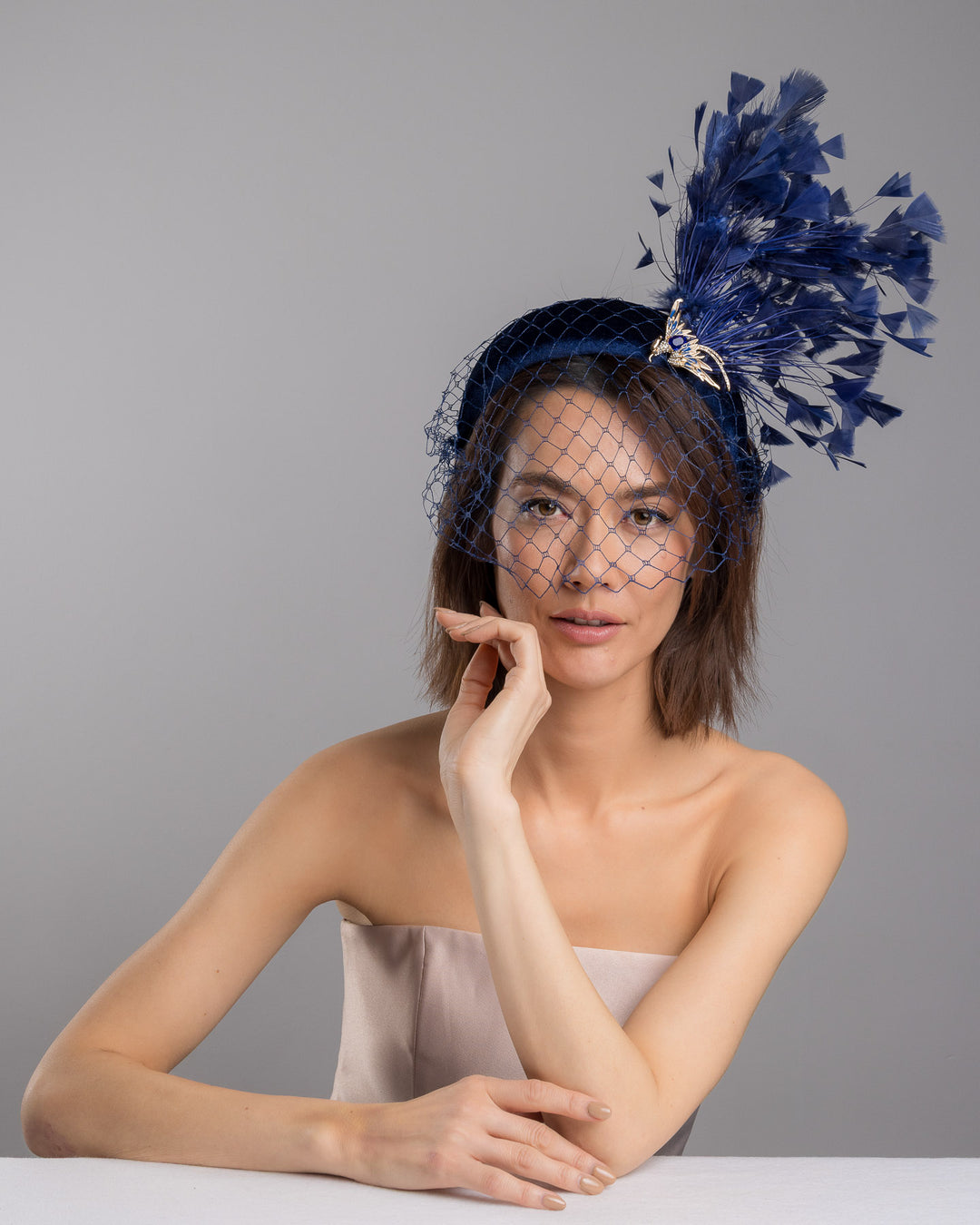 Female model is posing wearing Phoenix the navy coloured high velvet headband, covering half of the face with a stiffened navy veil elevated with a gold coloured metal bird with a large navy rhinestone and navy feathers behind the word placed high and like a fan. Made by Merve Bayindir Millinery.