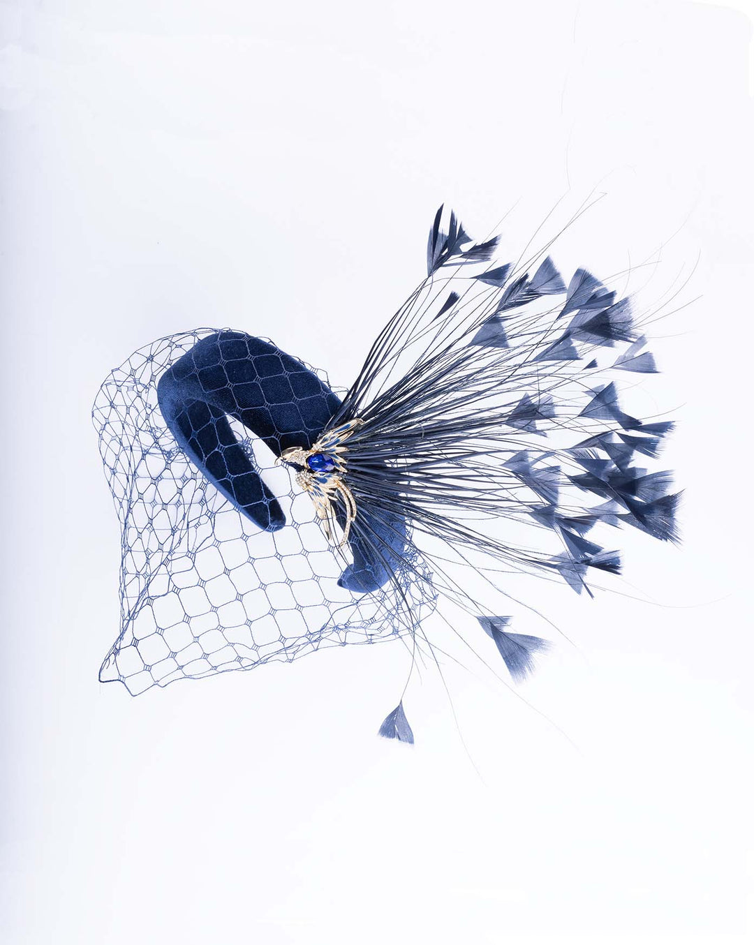 Still life picture of navy coloured high velvet headband, covering half of the face with a stiffened navy veil elevated with a gold coloured metal bird with a large navy rhinestone and navy feathers behind the word placed high and like a fan. Made by Merve Bayindir Millinery.