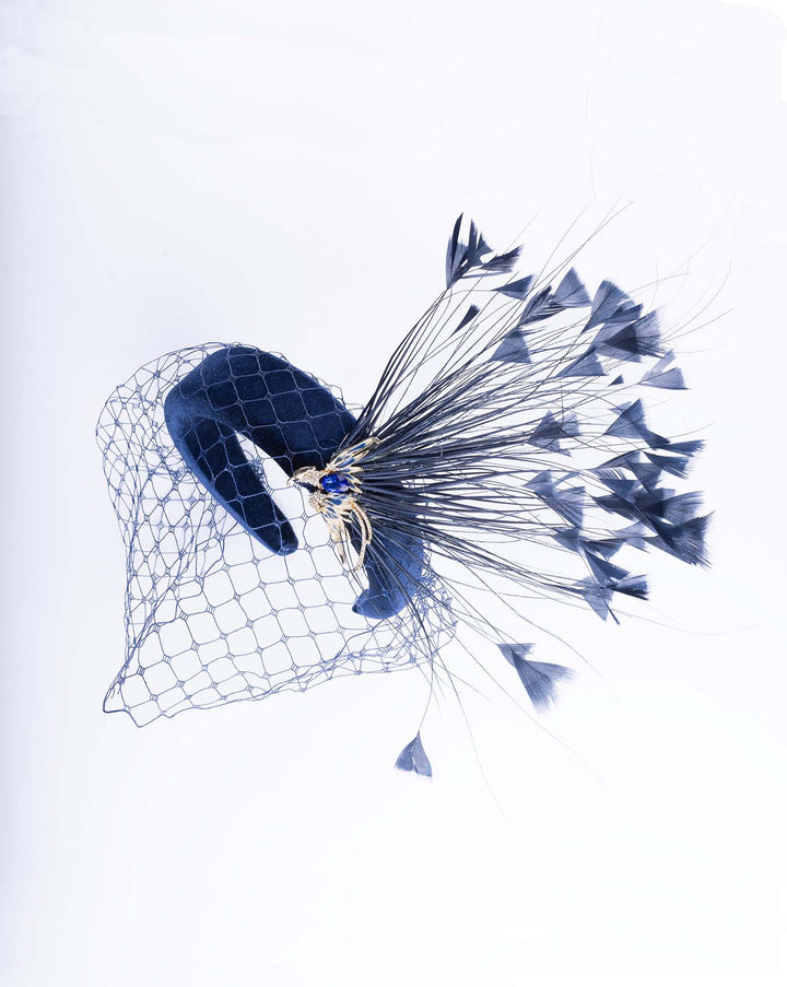 Still life picture of navy coloured high velvet headband, covering half of the face with a stiffened navy veil elevated with a gold coloured metal bird with a large navy rhinestone and navy feathers behind the word placed high and like a fan. Made by Merve Bayindir Millinery.