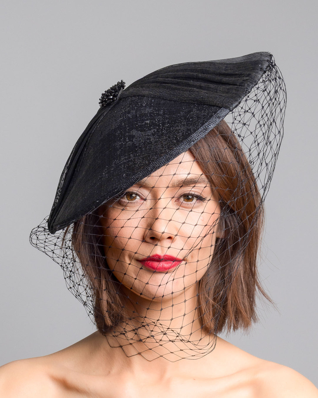 The model is showcasing the front side close up of the black oval fascinator crafted from fine sinamay, featuring soft pleated detailing across the crown, a small beaded accent at the top, and a delicate birdcage veil that drapes over the face. Designed and made by Merve Bayindir Millinery.
