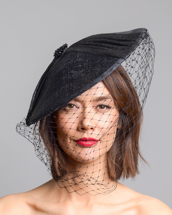 The model is showcasing the front side close up of the black oval fascinator crafted from fine sinamay, featuring soft pleated detailing across the crown, a small beaded accent at the top, and a delicate birdcage veil that drapes over the face. Designed and made by Merve Bayindir Millinery.
