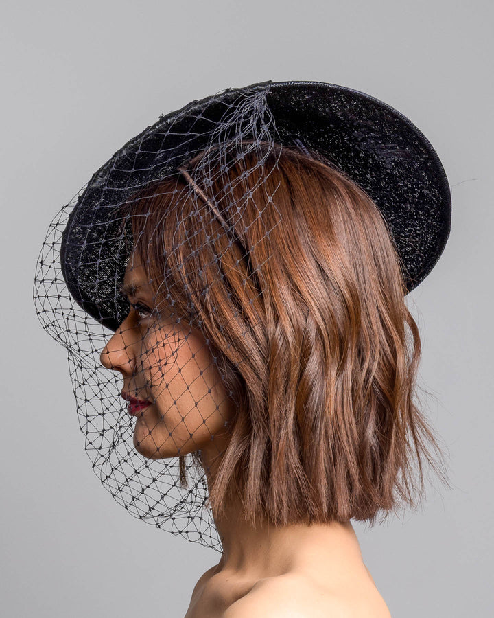 The model is showcasing the left side close up of the black oval fascinator crafted from fine sinamay, featuring soft pleated detailing across the crown, a small beaded accent at the top, and a delicate birdcage veil that drapes over the face. Designed and made by Merve Bayindir Millinery.
