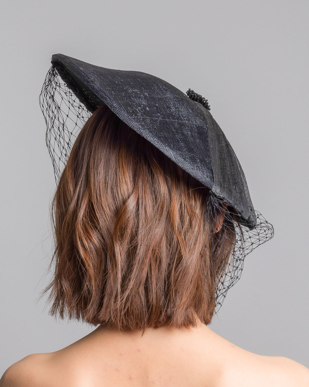 The model is showcasing the back side close up of the black oval fascinator crafted from fine sinamay, featuring soft pleated detailing across the crown, a small beaded accent at the top, and a delicate birdcage veil that drapes over the face. Designed and made by Merve Bayindir Millinery.
