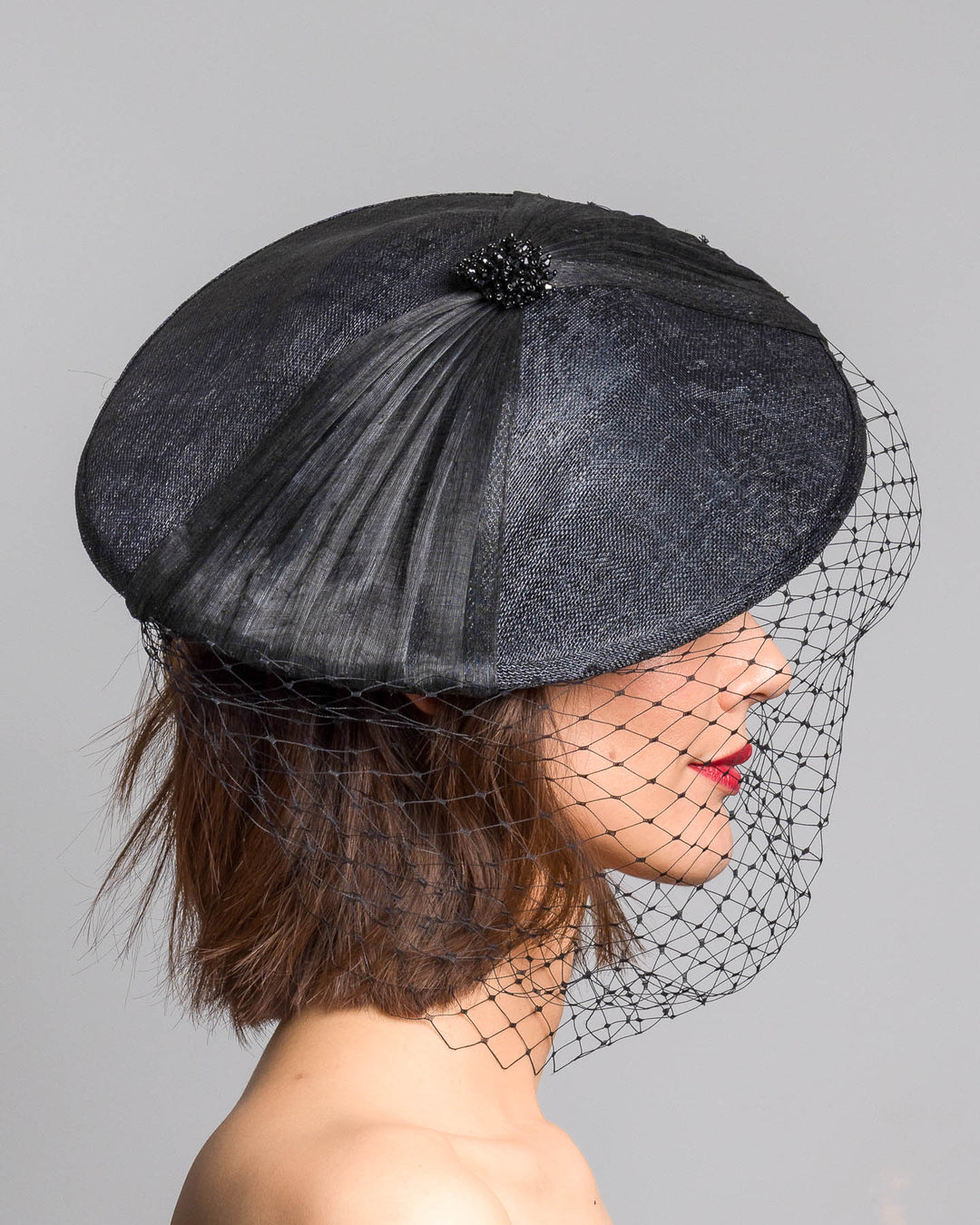 The model is showcasing the right side close up of the black oval fascinator crafted from fine sinamay, featuring soft pleated detailing across the crown, a small beaded accent at the top, and a delicate birdcage veil that drapes over the face. Designed and made by Merve Bayindir Millinery.
