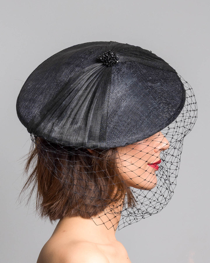 The model is showcasing the right side close up of the black oval fascinator crafted from fine sinamay, featuring soft pleated detailing across the crown, a small beaded accent at the top, and a delicate birdcage veil that drapes over the face. Designed and made by Merve Bayindir Millinery.
