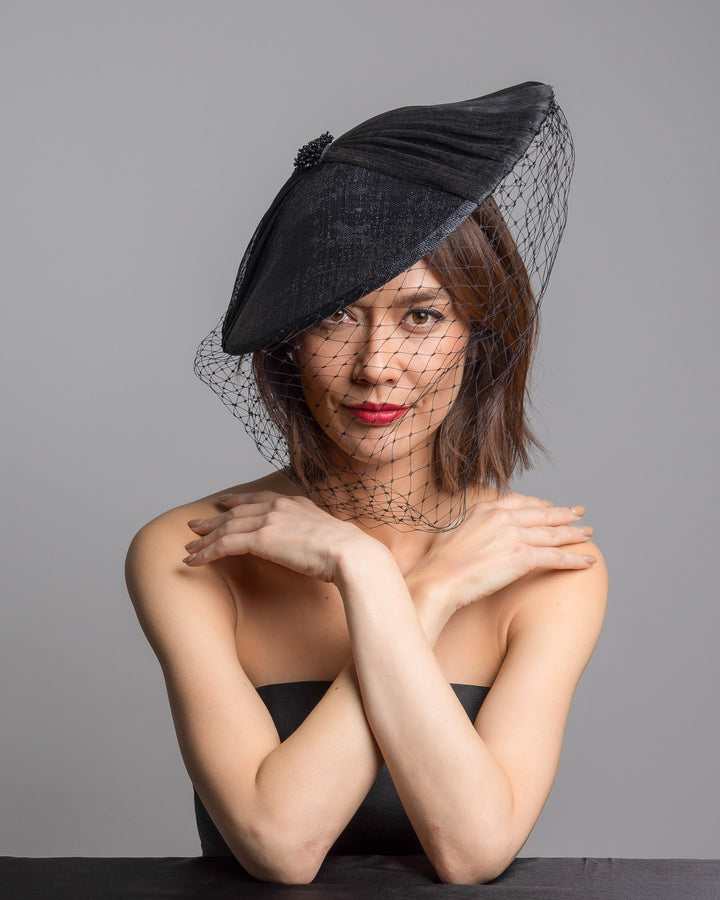 The female model is posing, wearing 
Ravenne, the black oval fascinator crafted from fine sinamay, featuring soft pleated detailing across the crown, a small beaded accent at the top, and a delicate birdcage veil that drapes over the face. Designed and made by Merve Bayindir Millinery.

