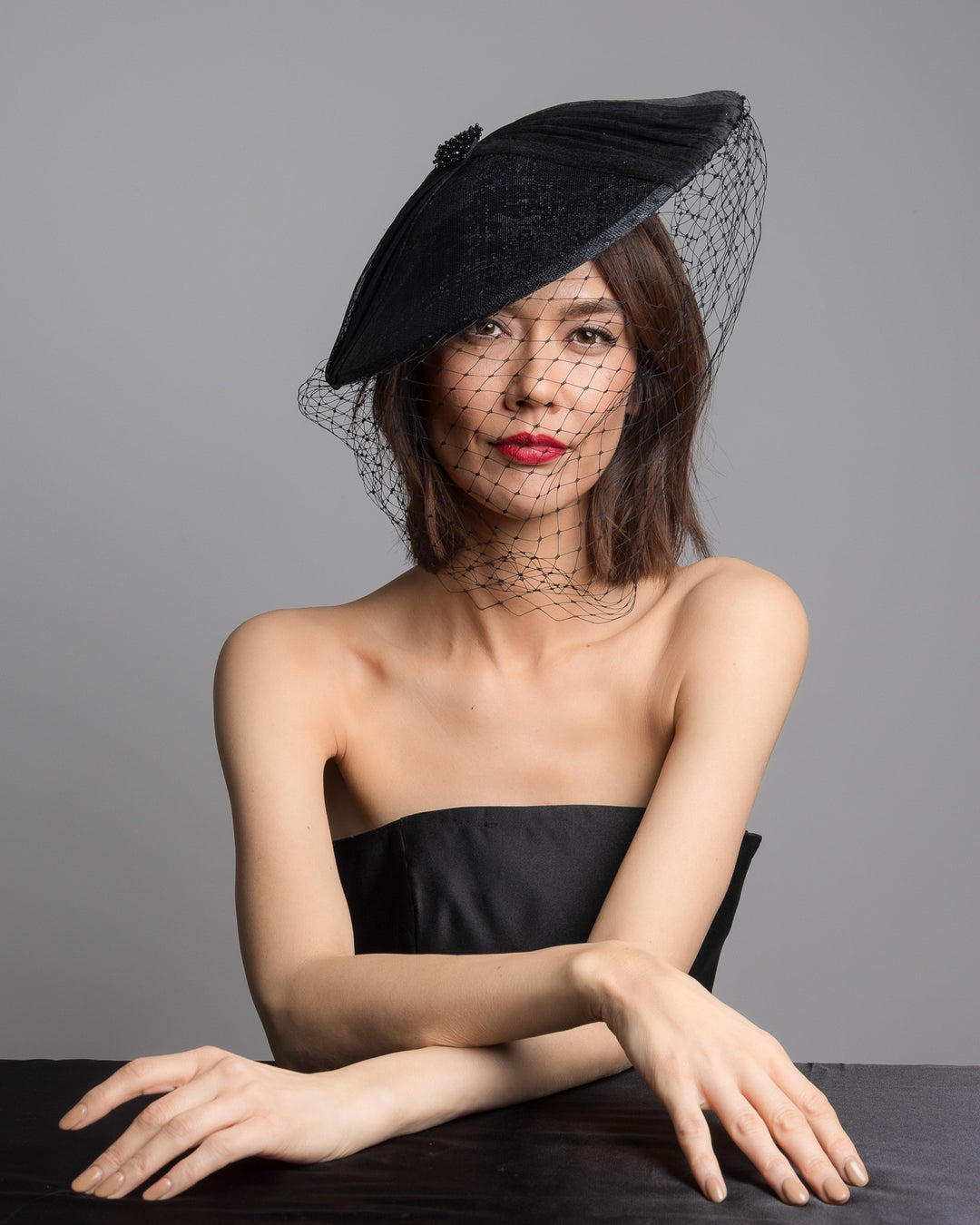 The female model is posing, wearing 
Ravenne, the black oval fascinator crafted from fine sinamay, featuring soft pleated detailing across the crown, a small beaded accent at the top, and a delicate birdcage veil that drapes over the face. Designed and made by Merve Bayindir Millinery.
