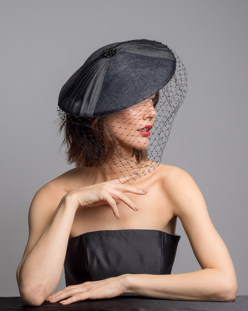 The female model is posing, wearing 
Ravenne, the black oval fascinator crafted from fine sinamay, featuring soft pleated detailing across the crown, a small beaded accent at the top, and a delicate birdcage veil that drapes over the face. Designed and made by Merve Bayindir Millinery.
