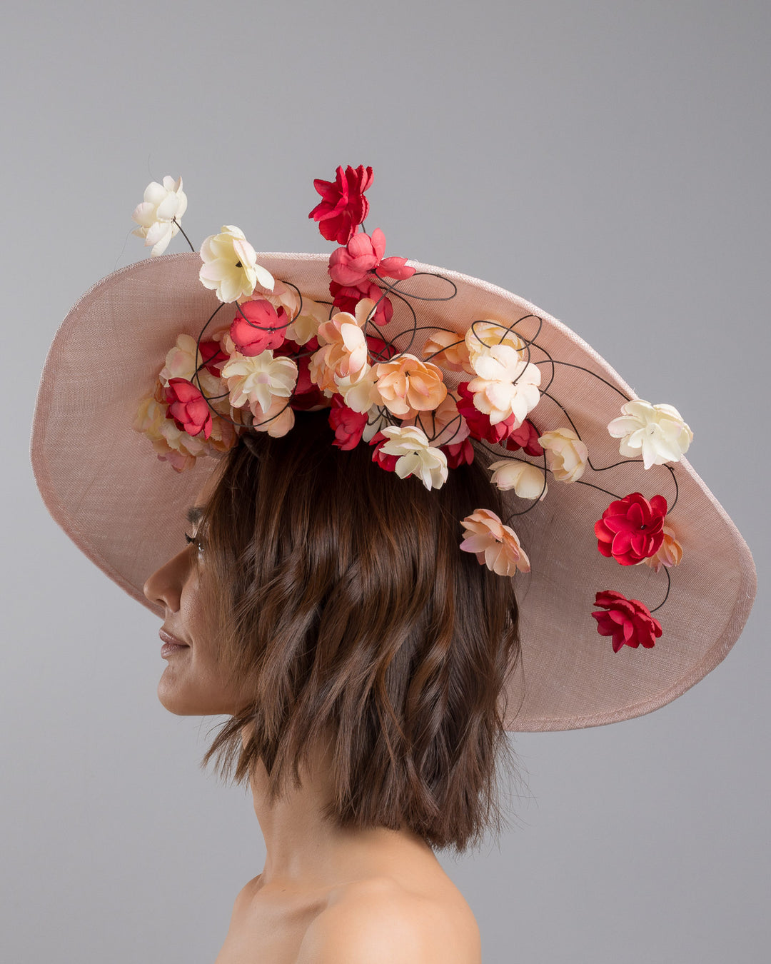 The model is showcasing the left side close up of the soft blush sinamay base hat with an elegant sculpted brim, adorned with cascading clusters of cream, peach, and deep red flowers on fine wired stems, creating a romantic, floating garden effect. Designed and made by Merve Bayindir Millinery.
