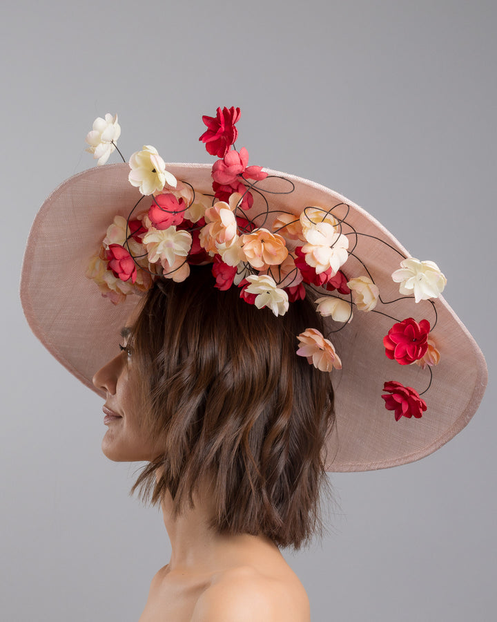 The model is showcasing the left side close up of the soft blush sinamay base hat with an elegant sculpted brim, adorned with cascading clusters of cream, peach, and deep red flowers on fine wired stems, creating a romantic, floating garden effect. Designed and made by Merve Bayindir Millinery.
