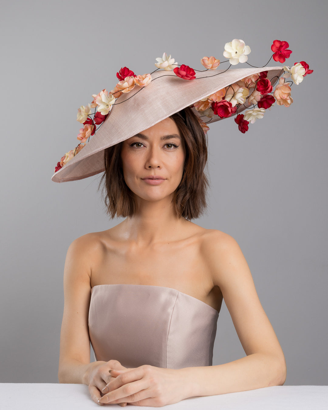 The female model is posing, wearing
Rivane, the soft blush sinamay base hat with an elegant sculpted brim, adorned with cascading clusters of cream, peach, and deep red flowers on fine wired stems, creating a romantic, floating garden effect. Designed and made by Merve Bayindir Millinery.
