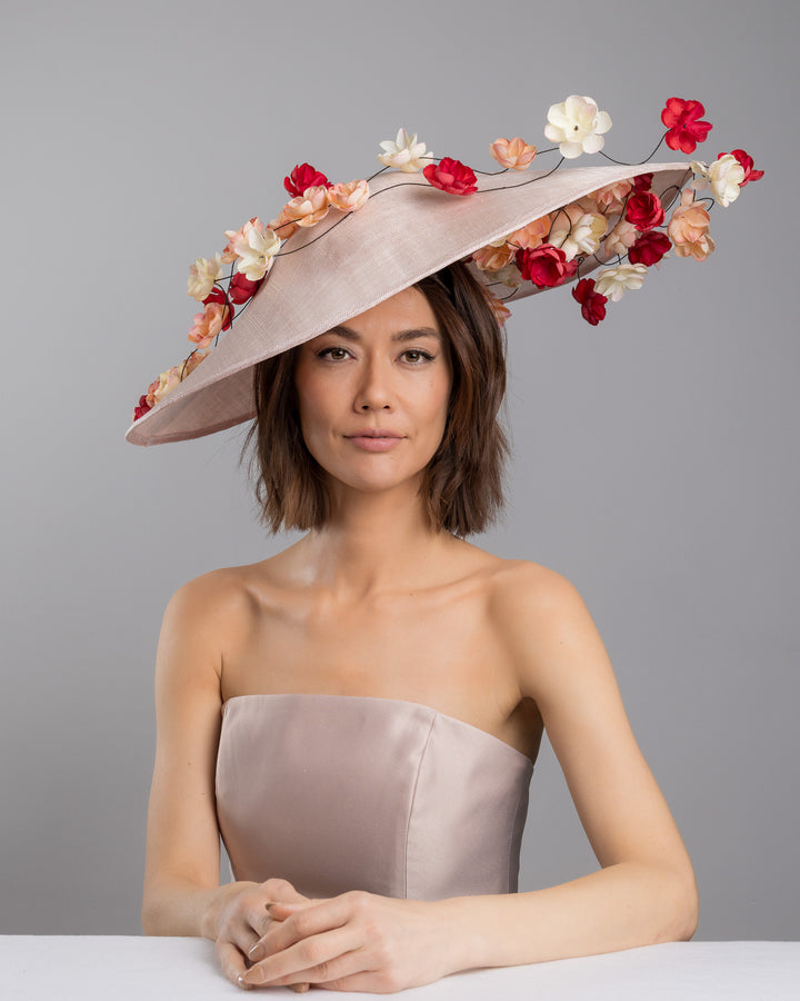 The female model is posing, wearing
Rivane, the soft blush sinamay base hat with an elegant sculpted brim, adorned with cascading clusters of cream, peach, and deep red flowers on fine wired stems, creating a romantic, floating garden effect. Designed and made by Merve Bayindir Millinery.
