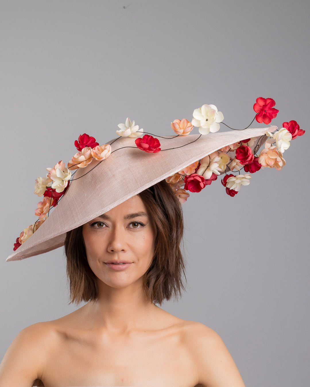 The model is showcasing the front side close up of the soft blush sinamay base hat with an elegant sculpted brim, adorned with cascading clusters of cream, peach, and deep red flowers on fine wired stems, creating a romantic, floating garden effect. Designed and made by Merve Bayindir Millinery.
