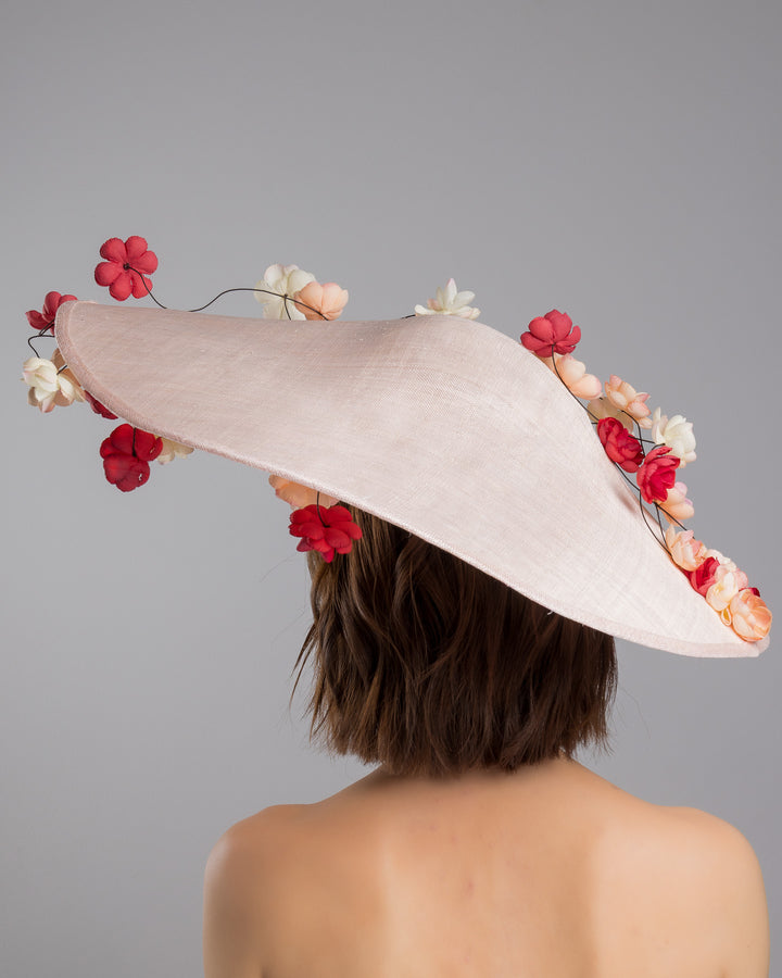 The model is showcasing the back side close up of the soft blush sinamay base hat with an elegant sculpted brim, adorned with cascading clusters of cream, peach, and deep red flowers on fine wired stems, creating a romantic, floating garden effect. Designed and made by Merve Bayindir Millinery.
