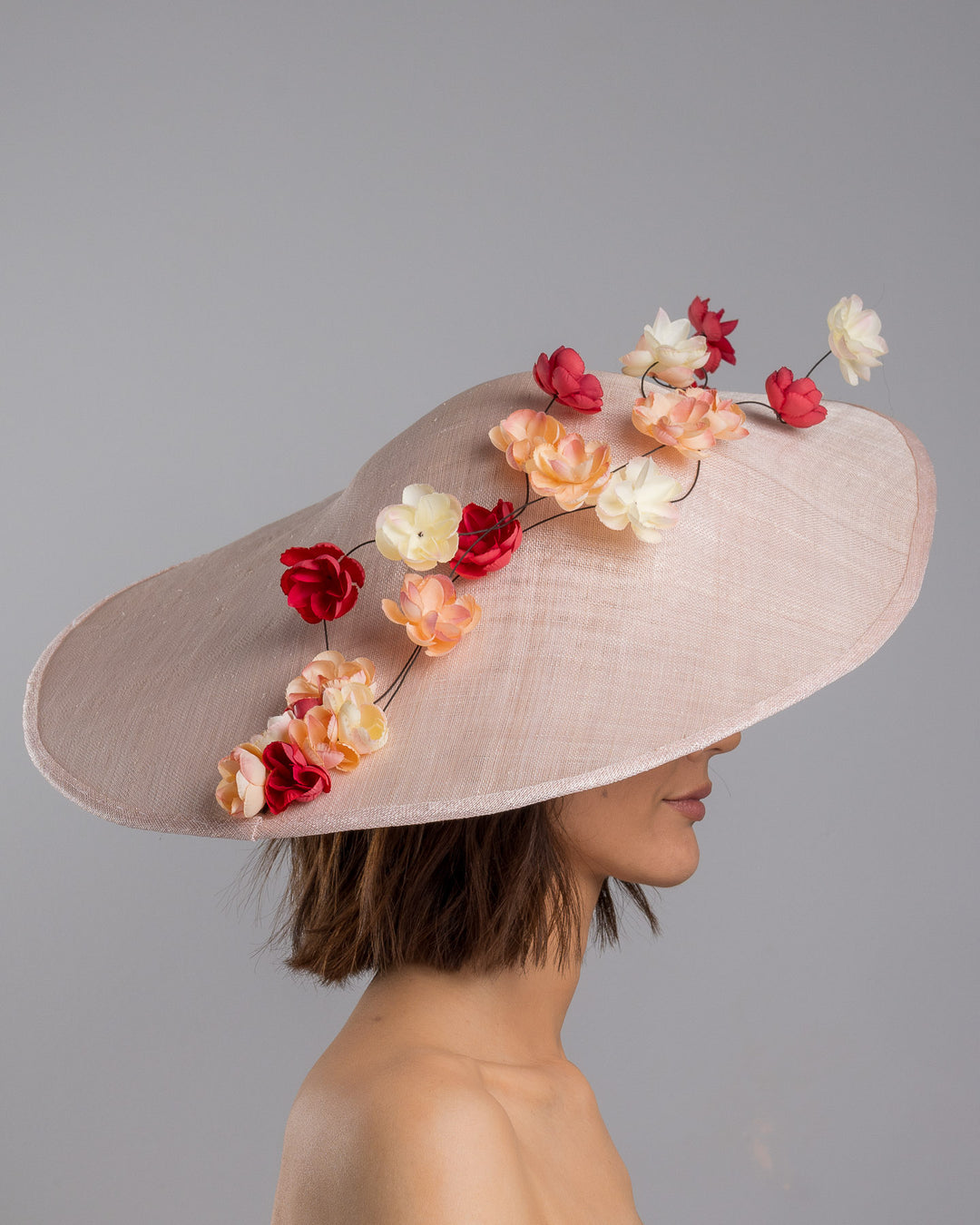 The model is showcasing the right side close up of the soft blush sinamay base hat with an elegant sculpted brim, adorned with cascading clusters of cream, peach, and deep red flowers on fine wired stems, creating a romantic, floating garden effect. Designed and made by Merve Bayindir Millinery.

