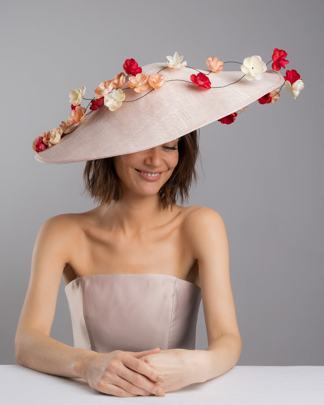The female model is posing, wearing
Rivane, the soft blush sinamay base hat with an elegant sculpted brim, adorned with cascading clusters of cream, peach, and deep red flowers on fine wired stems, creating a romantic, floating garden effect. Designed and made by Merve Bayindir Millinery.
